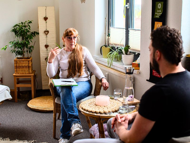 Heidi sitzt mit einem Klemmbrett in einem Korbsessel und führt ein aufmerksames Vorgespräch zur Energiearbeit mit einem männlichen Klienten.