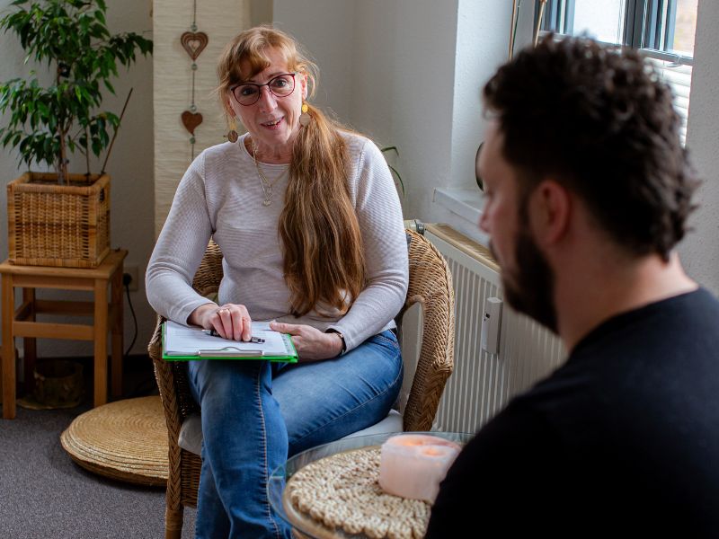 Heidi Siegert sitzt mit einem Klemmbrett auf dem Schoß in einem Korbsessel und bespricht Details zur Ernährungsberatung in Bautzen mit einem Klienten.