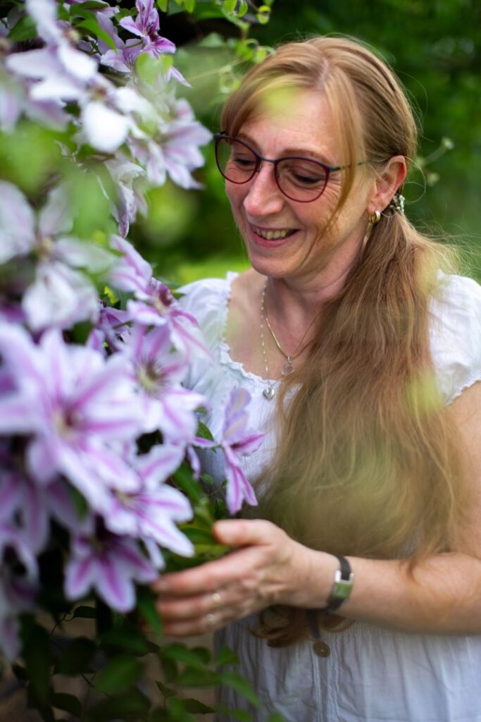 Heidi Siegert steht lächelnd im Grünen neben einer Pflanze mit großen violetten Blüten und strahlt die Frische aus für das energetische Facelifting in Bautzen.