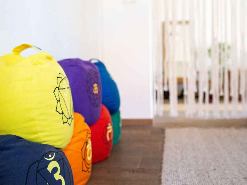 Mehrere bunte Kissen mit aufgedruckten Chakra Symbolen liegen auf dem Fußboden vor einem Fadenvorhang im Raum für das energetische Facelifting in Bautzen.
