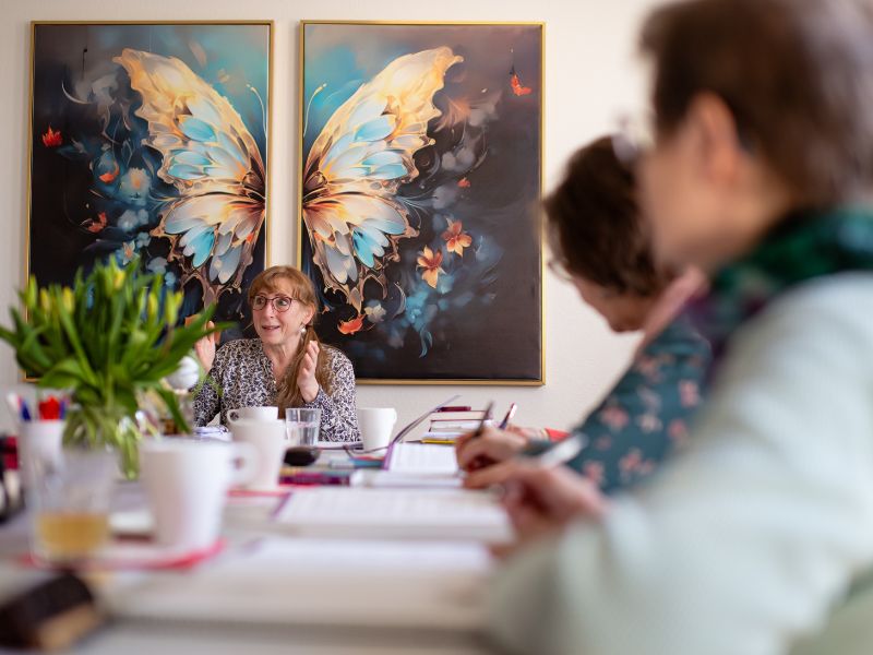 Heidi Siegert sitzt an einem großen Tisch als Dozentin und unterrichtet eine Gruppe vor einem großen Schmetterlingsgemälde passend zur Pranaheilung in Bautzen.